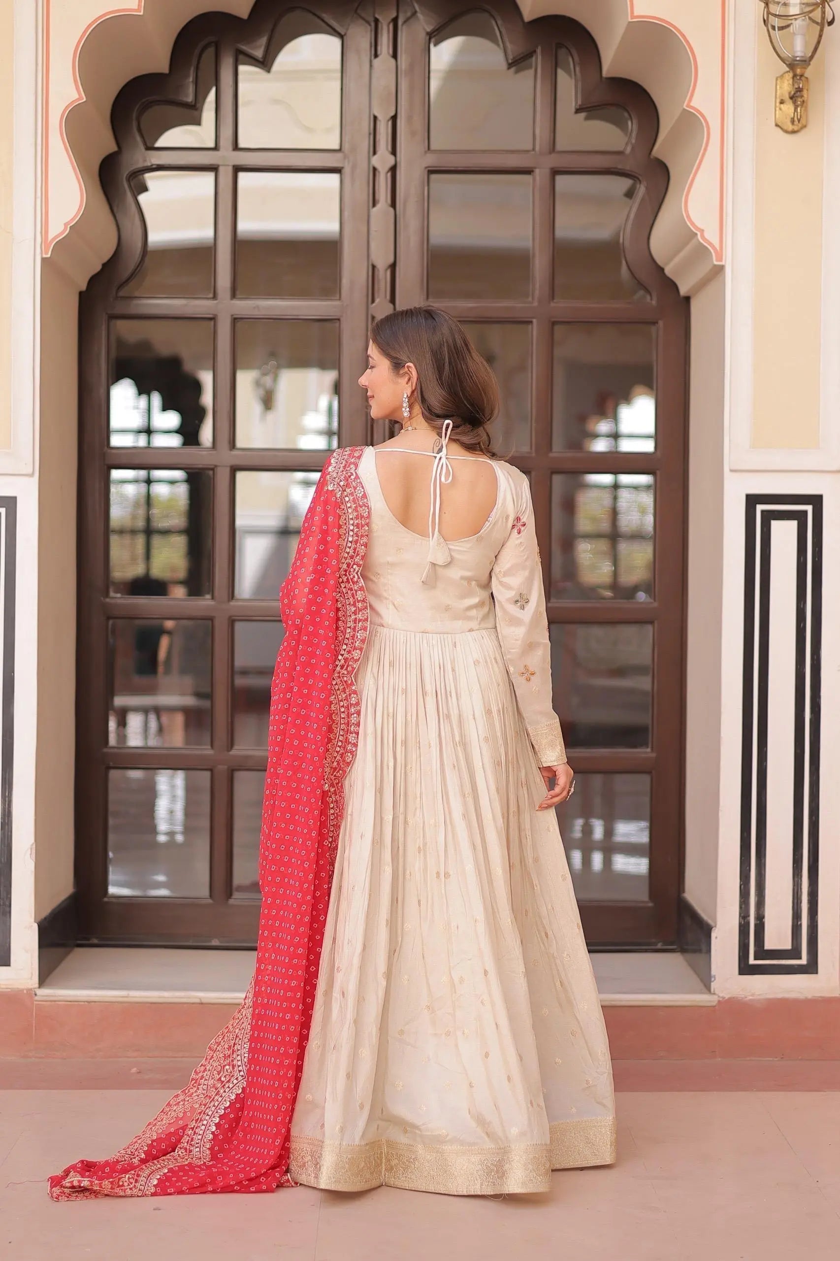 Woman in a cream traditional outfit with a red dupatta standing in front of an ornate doorway.